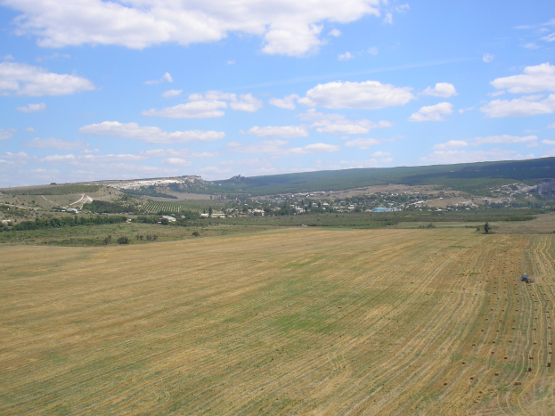 Ein Traktor, der auf einem bewirtschafteten Feld mit Bäumen, Gebäuden, Hügeln und einem bewölkten Himmel im Hintergrund arbeitet.