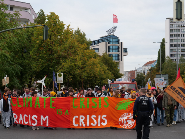 Eine Gruppe von Menschen marschiert eine baumbestandene Straße entlang und hält ein Banner mit der Aufschrift "Klimakrise ist eine Krise", mit parkenden Fahrzeugen, Gebäuden und einem klaren blauen Himmel im Hintergrund.
