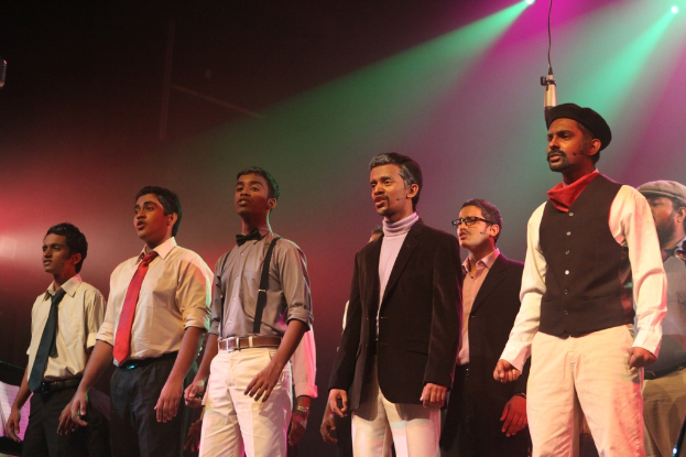 Eine Gruppe von Männern auf einer Bühne, die ein Drama aufführen, mit grünen Scheinwerfern dahinter.