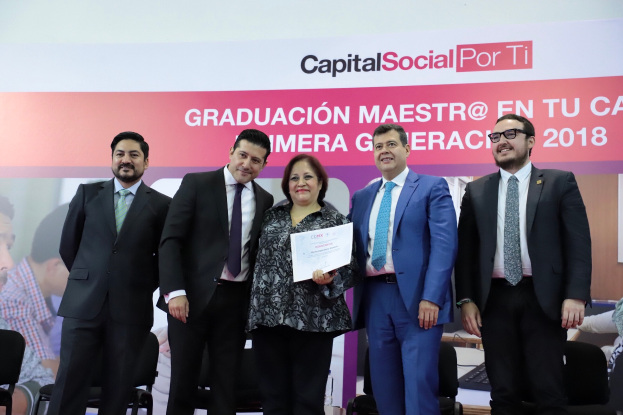 Eine Gruppe von Menschen vor einem Bildschirm, mit einer Frau in der Mitte, die eine Urkunde hält, und anderen rechts sitzend, mit einem Banner im Hintergrund mit Text und Bildern von Menschen.