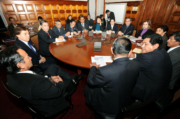 Eine Runde-Tisch-Besprechung mit Delegierten, die diskutieren, umgeben von Mikrofonen, Papieren, Kabeln und Dateien, mit einer hölzernen Wand im Hintergrund.