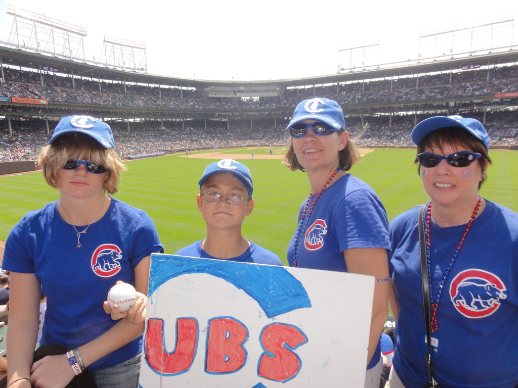 Drei Frauen in Kappen und Brillen, ein Junge in Brille und Kappe mit einem Ball und einer Tafel, die in einem Stadion mit Publikum und Feld hinter ihnen stehen, unter einem Himmel mit wenigen Lichtern.