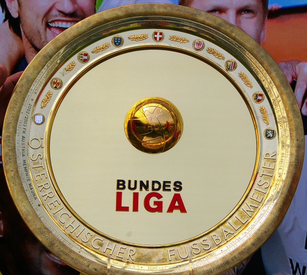 Ein Mann, der ein goldenes Schild mit der Aufschrift 'Bundesliga' hält, mit einem Banner im Hintergrund, auf dem drei Personen zu sehen sind.