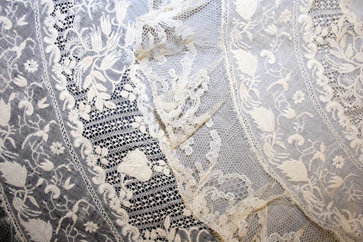 Nahaufnahme eines weißen Hochzeitskleids mit zarter, kunstvoller Spitzenverzierung in verschiedenen Mustern auf einem leichten, luftigen Stoff.