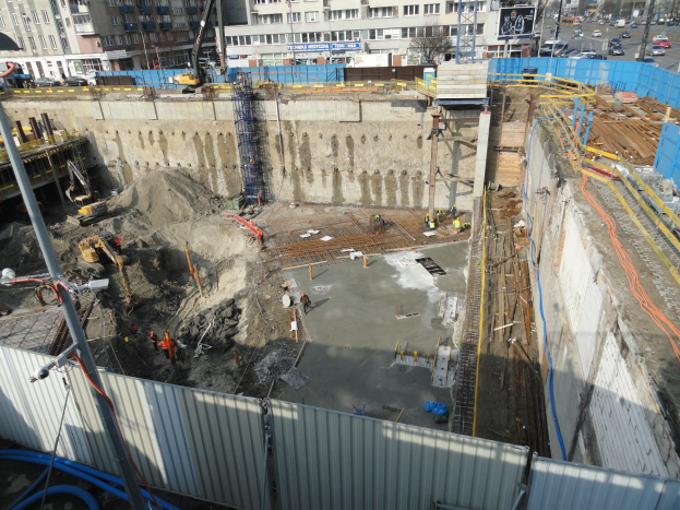 Eine Baustelle mit einem großen Haufen Erde in der Mitte, umgeben von Gebäuden, Bäumen, Pfählen, Fahrzeugen und anderen Gegenständen, mit einem Metallblech und Rohren unten.