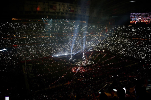 Große Menschenmenge in einem hell erleuchteten Stadion bei Nacht mit einem Bildschirm, der links die Ergebnisse des Super Bowl XLVII-Halbtzeitshows anzeigt.