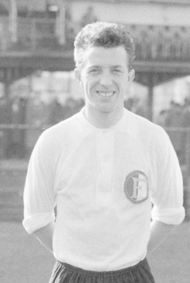Ein Schwarz-Weiß-Foto eines grinsenden Mannes in T-Shirt und Shorts auf einem Fußballfeld mit einem Gebäude im Hintergrund, der mutmaßlich Mario Mandzukic ist.