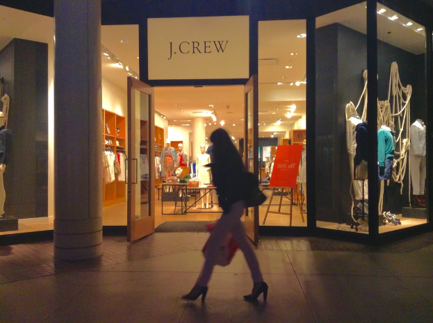 Ein Einkaufszentrum mit Glaswänden und -türen, das Schaufensterpuppen, Tische, hängende Kleider, Schränke und eine Frau zeigt, die daneben geht.