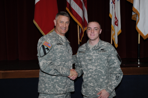 Zwei Personen in Uniformen geben sich die Hand und lächeln, vor vier Fahnen auf Stangen vor dunklem Hintergrund.