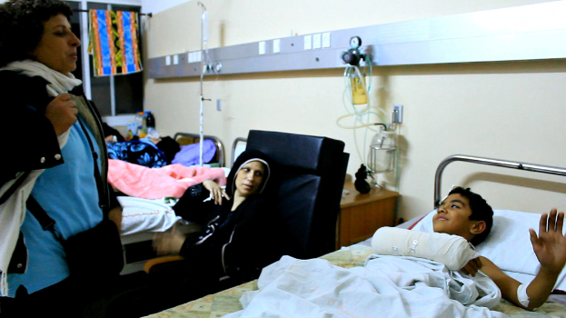 Ein Krankenhauszimmer mit mehreren Betten, an denen Patienten liegen, sowie Infusionsflaschen auf Ständern zwischen den Betten und eine Frau mit einer Tasche in der Nähe eines Patienten.