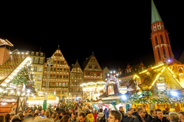 Ein lebendiger Weihnachtsmarkt in Nürnberg, Deutschland, mit Menschen um beleuchtete und geschmückte Stände, Gebäuden mit Fenstern und einem Kirchturm im Hintergrund unter einem dunklen Himmel.