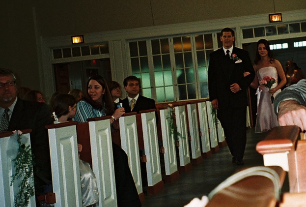 Eine Gruppe von Menschen sitzt auf einer Bank, während eine Braut und ein Bräutigam den Weg zu ihnen entlanggehen, die Braut hält einen Blumenstrauß, mit Laternen und einem Fenster im Hintergrund, was auf eine Kirchenatmosphäre hindeutet.