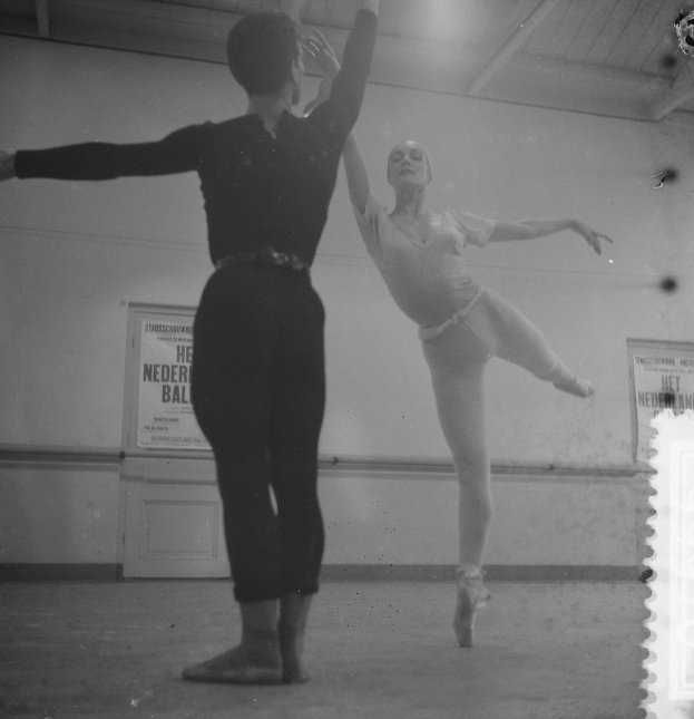 Ein Schwarz-Weiß-Foto von einem Mann und einer Frau, die in einem Studio tanzen, wobei die Frau in der Luft ist und eine mit Plakaten bedeckte Wand im Hintergrund zu sehen ist, die von Deckenlampen beleuchtet wird.