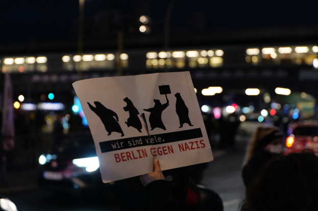 Eine Person mit einem Schild mit der Aufschrift "Berlin gegen Nazis" vor einer Menge und Fahrzeugen auf einer Straße, mit einem beleuchteten Gebäude im Hintergrund.