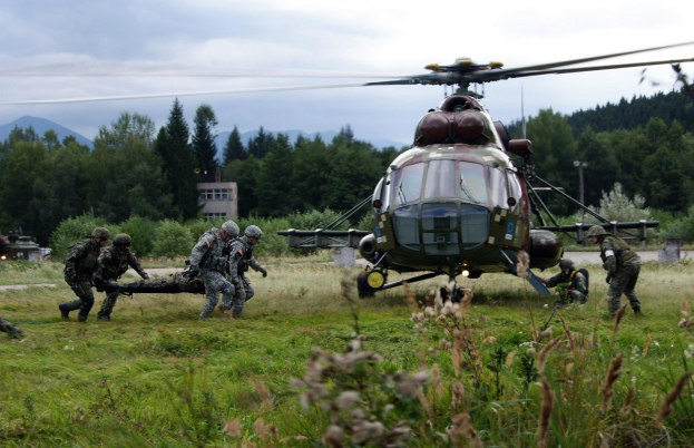 Soldaten tragen einen Mann auf einer Trage vor einem Hubschrauber, umgeben von Gras und Pflanzen, mit Bäumen, Gebäuden, Hügeln und einem bewölkten Himmel im Hintergrund.
