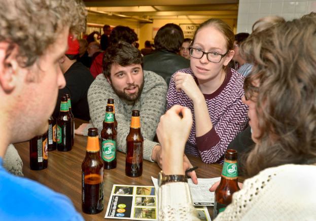 Mehrere Personen stehen auf beiden Seiten eines Tisches mit Bierflaschen, der wie eine Bar aussieht.