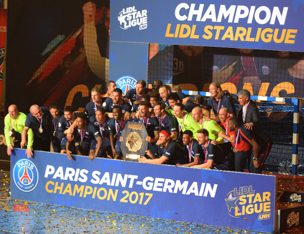 Paris Saint-Germain-Spieler feiern mit der Ligue-1-Trophäe, tragen Medaillen, vor einer Tafel mit Text und Logos, mit einem Netz auf der rechten Seite und einem Bildschirm im Hintergrund.