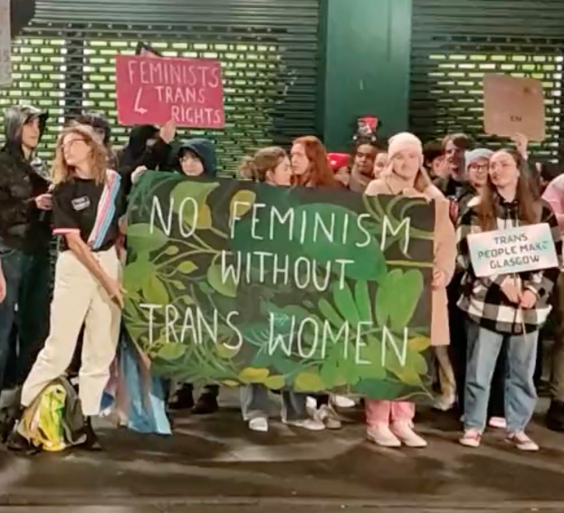 Eine Gruppe von Menschen hält ein Transparent mit der Aufschrift 'Kein Feminismus ohne Trans-Frauen' und verschiedene Schilder und steht an einem öffentlichen Ort.