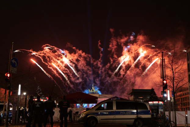 Polizeifahrzeug vor Menschenmenge mit beleuchteten Gebäuden, Bäumen, Polen, Verkehrsampeln, Schildern und Zelten im Hintergrund bei farbigen Feuerwerken.