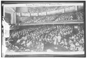 Ein Schwarz-Weiß-Foto einer großen Menschenmenge in einem Gebäude, einige sitzen, einige stehen, Schilder mit Text und eine Wand im Hintergrund. Unten steht der Text "Wahlberechtigte Frauen gründen eine Frauenpartei".