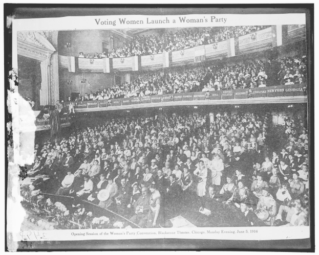 Ein Schwarz-Weiß-Foto einer großen Menschenmenge in einem Gebäude, einige sitzen, einige stehen, Schilder mit Text und eine Wand im Hintergrund. Unten steht der Text "Wahlberechtigte Frauen gründen eine Frauenpartei".