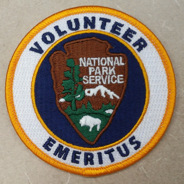 Ein rundes Patch mit einem blauen Rand und einem weißen Hintergrund, das schwarze Text 'Volunteer National Park Service Emeritus' in der Mitte eines gelben Kreises zeigt.