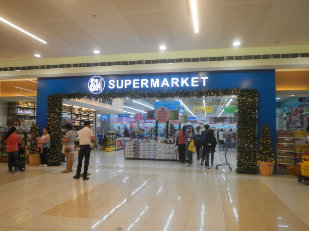 Eine Gruppe von Menschen vor einem Supermarkt, einige mit Einkaufstägern, einem Einkaufswagen, Topfpflanze, Regalen, Schildern und Deckenleuchten.