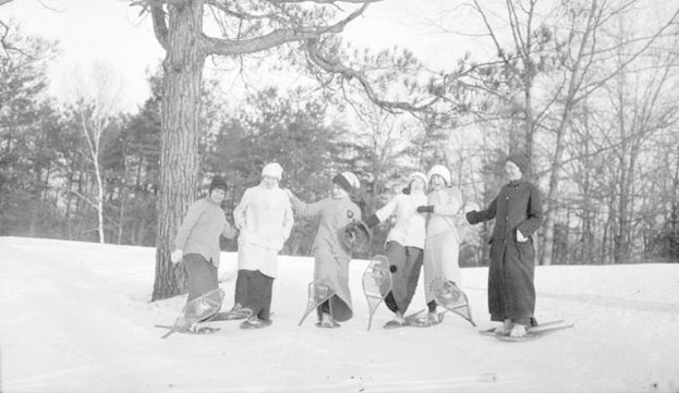 Sechs Frauen auf Snowboards, positioniert auf einer schneebedeckten Fläche mit einem Hintergrund aus Bäumen.