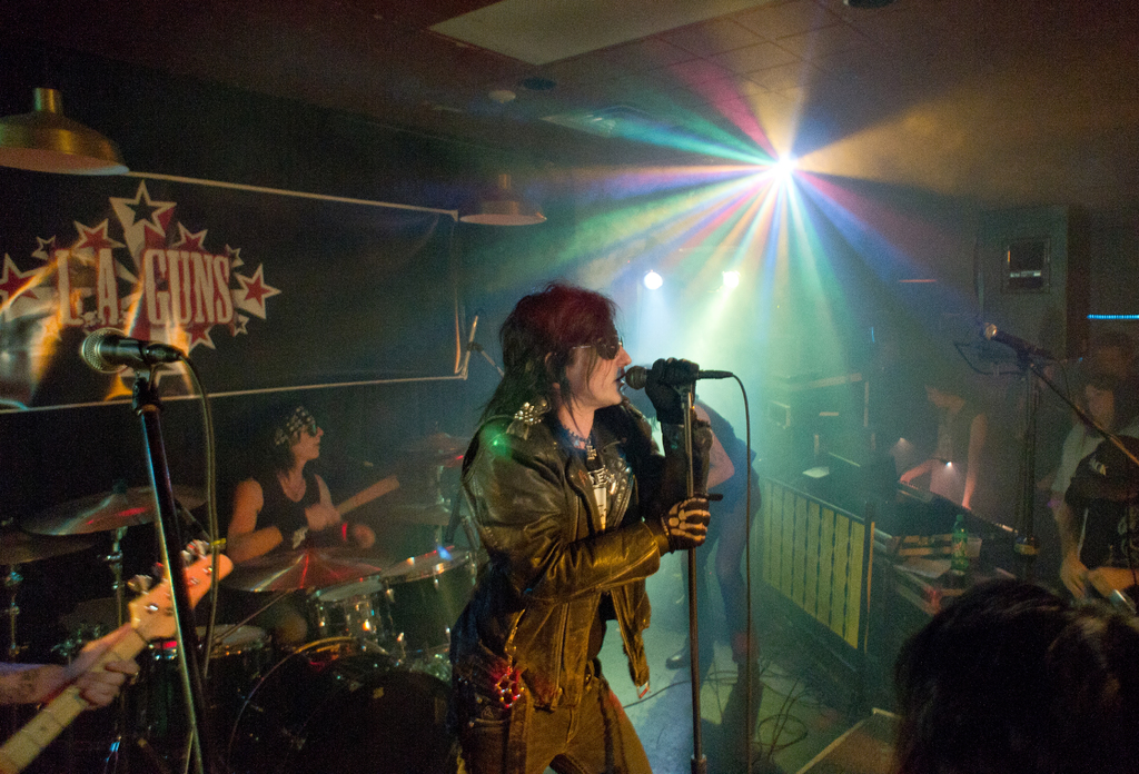 Eine Frau in einer schwarzen Jacke singt in ein Mikrofon auf der Bühne, mit einem Mann in Sonnenbrille und einem Headband, der hinter ihr ein Instrument spielt, und einem Plakat mit 'LA Guns' im Hintergrund.
