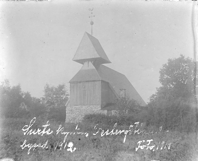 Schwarzes und weißes Foto einer Kirche mit einem Kirchturm, umgeben von Bäumen und Pflanzen, unter einem bewölkten Himmel, beschriftet mit "Kirche des Heiligen Grabes" unten.