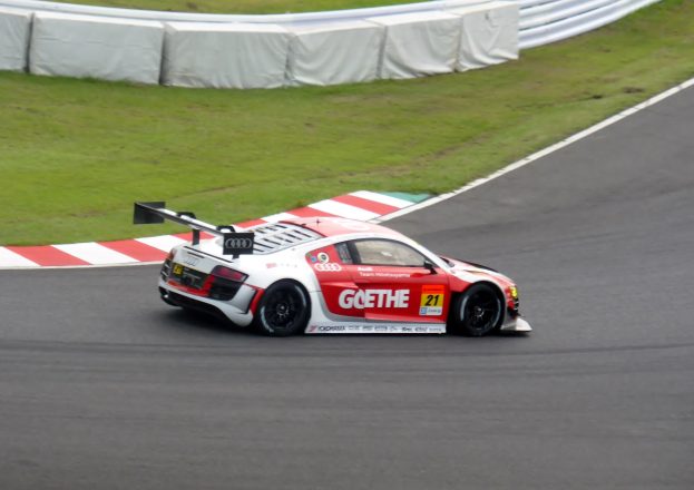 Ein leuchtend roter und weißer Audi R8 LMS GT3 Rennwagen auf einer Rennstrecke, umgeben von grünem Gras und einer Umrandung im Hintergrund.