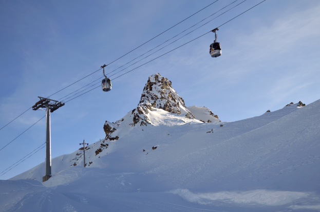 Skilift fährt einen schneebedeckten Berg hinauf mit Seilbahnkabinen im Vordergrund, verbunden durch Pfosten und Drähte, vor einem bewölkten Himmel.