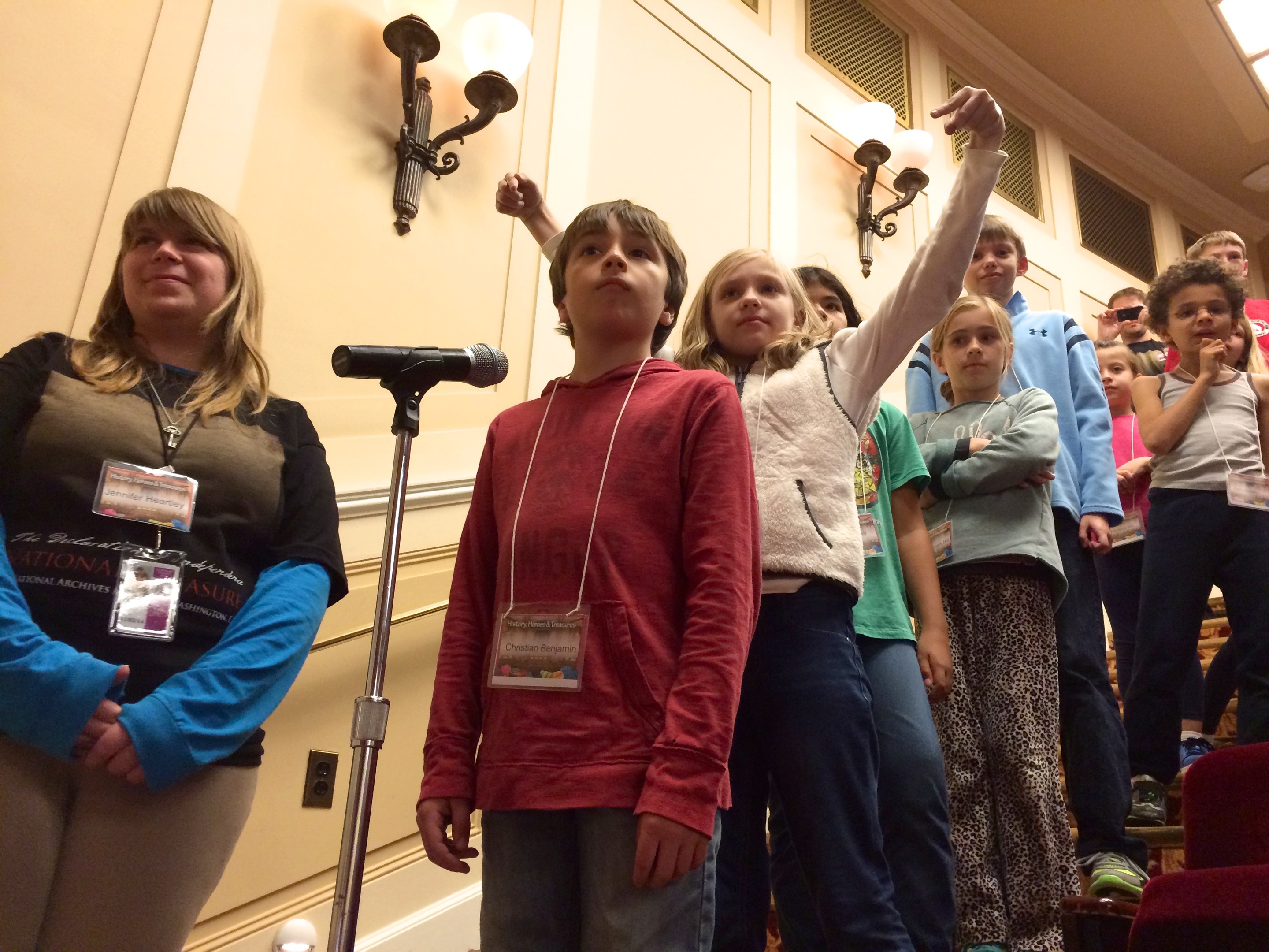 Kinder singen auf einer Nationalversammlung, begleitet von einer Frau, mit Stühlen und beleuchteter Wand im Hintergrund.
