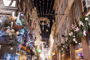 Ein geschäftiger Weihnachtsmarkt in Straßburg, Frankreich, mit Menschen, die zwischen festlichen Dekorationen wie Weihnachtsbäumen und Lichtern auf geschmückten Gebäuden unter einem sichtbaren Himmel spazieren.