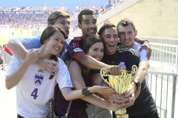 Sechs Personen, darunter zwei Frauen und vier Männer, halten einen Pokal und lächeln für ein Foto vor einem Eisengeländer, mit einer Wand und einem Geländer oben rechts und Menschen und Lichtern im Hintergrund, möglicherweise in einem Stadion.