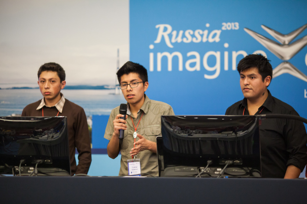 Drei Jungen stehen neben einem Schreibtisch mit zwei Computersystemen, einer spricht in ein Mikrofon und trägt ID-Karten mit roten Markierungen, vor einer Wand mit einer Werbetafel, auf der 'Russia Imagine 2013' steht.