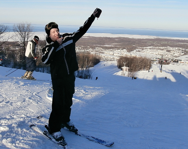Eine Person auf einem Snowboard in der Mitte des Bildes, umgeben von anderen Skifahrern, Bäumen, einem Pfahl, Wasser und Himmel im Hintergrund.