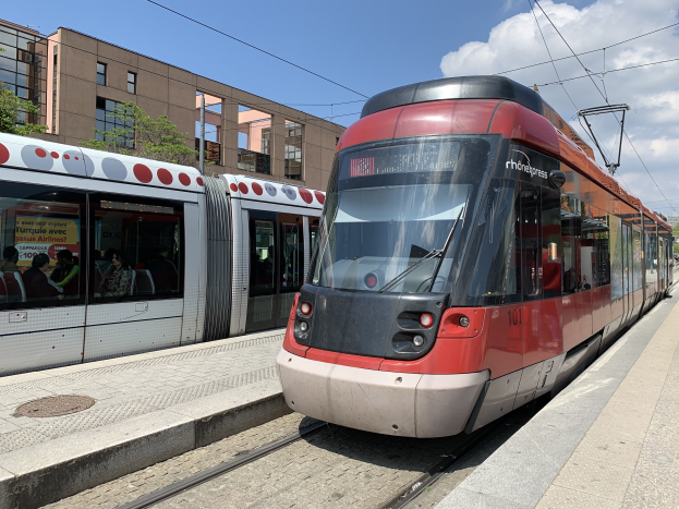 Ein roter und weißer Zug fährt auf Schienen neben einem Bahnsteig mit einigen Menschen darauf, mit Gebäuden, Bäumen, Pfählen und Drähten im Hintergrund, die das neue Straßenbahnsystem in Madrid darstellen.