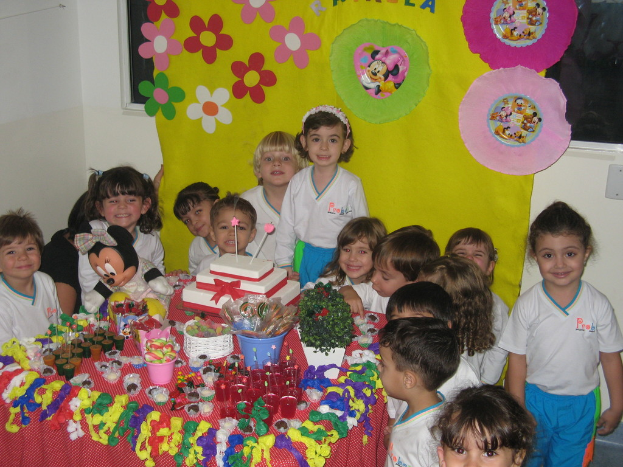 Eine Gruppe von Kindern sitzt um einen Tisch, der mit einem Kuchen, Tassen, Saft, Cremes, Löffeln, Eimern, Schokoladen, Lutschern, Ballons und einem Tuch gefüllt ist, mit einem gelben Tuch im Hintergrund neben einem Fenster.