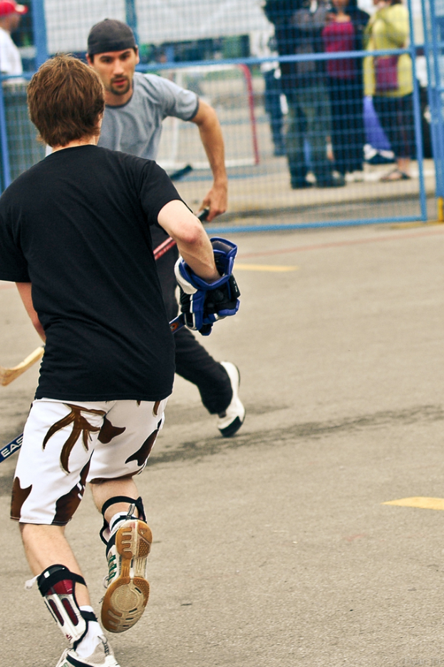 Zwei Personen spielen Hockey auf einer Straße mit einem Zaun dahinter und einer Menge Zuschauer im Hintergrund.