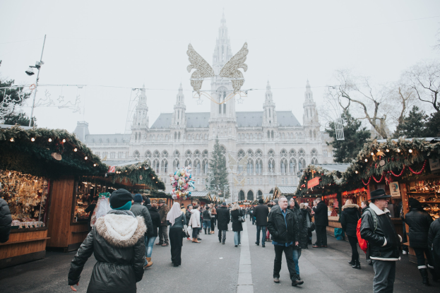 Ein geschäftiges Weihnachtsmarkt in Wien, Österreich mit Menschen, die zwischen Ständen mit Lichtern und festlichen Artikeln spazieren, vor einem Gebäude, Bäumen und einem klaren blauen Himmel.