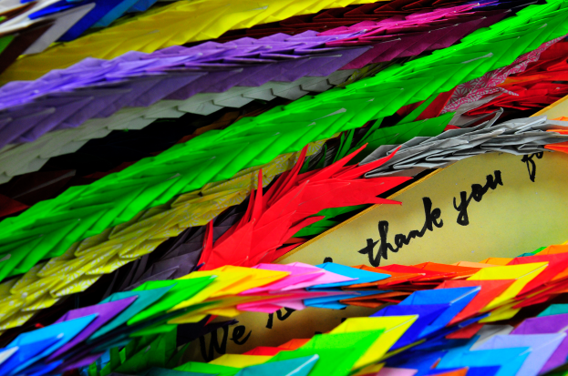 Ein Stapel bunter Origami-Kraniche in Rot, Blau, Gelb, Grün und Lila mit einer Dankeskarte obenauf.