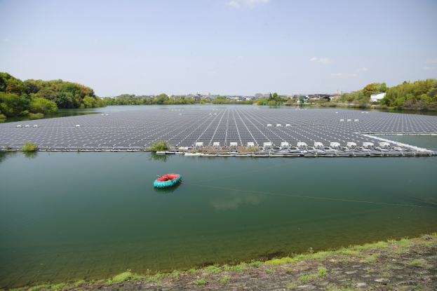 Ein kleines Boot treibt auf einem Gewässer, das von Grün umgeben ist, mit Solarpanelen auf der Wasseroberfläche und Gebäuden im Hintergrund unter einem klaren blauen Himmel.