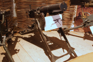 Eine Gatling Gun, eine Art Maschinengewehr, das von der britischen Armee während des Zweiten Weltkriegs verwendet wurde, wird auf einem Holzfußboden in einem Museum ausgestellt, mit einer Wand im Hintergrund und einer Texttafel auf der rechten Seite des Bildes.