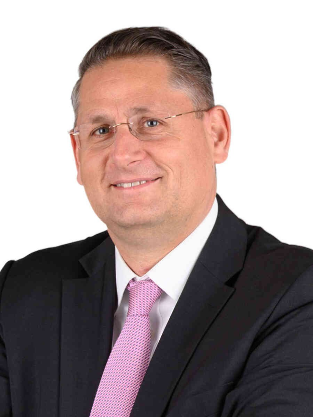 Ein lächelnder Mann im Anzug und Krawatte, der eine Brille trägt, vor einem weißen Hintergrund.