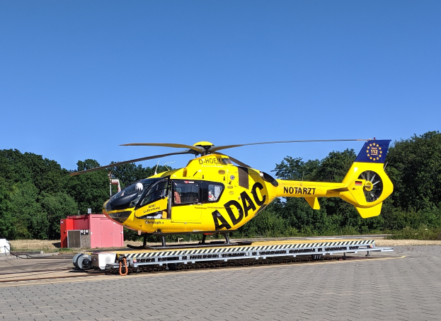 Ein gelber Hubschrauber steht auf dem Rollfeld eines Flughafens umgeben von Bäumen und einem klaren blauen Himmel, mit verschiedenen Gegenständen, die mit dem Hubschrauber in Verbindung stehen, verstreut.