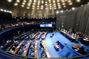 Eine Konferenzbühne mit sitzenden Personen, einem Tisch mit Papieren und Gegenständen, Fahnen und einer Skulptur im Hintergrund, mit Zuschauern an Tischen und hinter einer Absperrung, beleuchtet von Deckenlampen.
