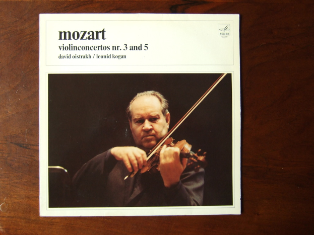 Ein Mann in einem schwarzen Mantel spielt Geige auf einem Holztisch, mit einem schwarzen Hintergrund und einer kleinen Inschrift über dem Bild.