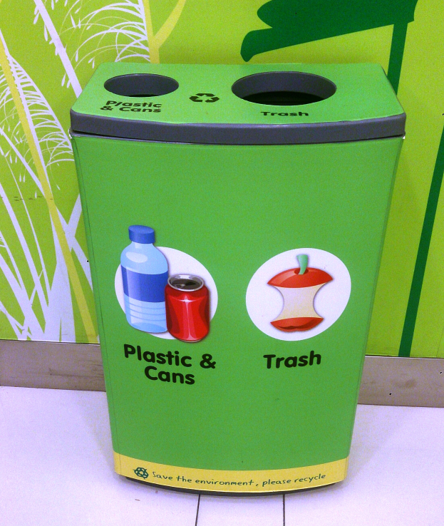 Ein grüner Mülltonne aus Kunststoff und Dosen auf einem weißen Boden steht, mit einem Plakat im Hintergrund.