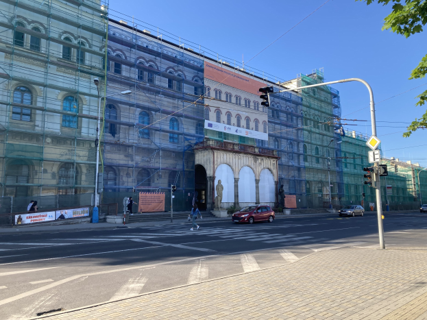 Großes Gebäude mit Gerüst, das renoviert wird, umgeben von Fahrzeugen, Fußgängern, Ampeln, Straßenschildern, Polen, Bäumen und einem klaren blauen Himmel.